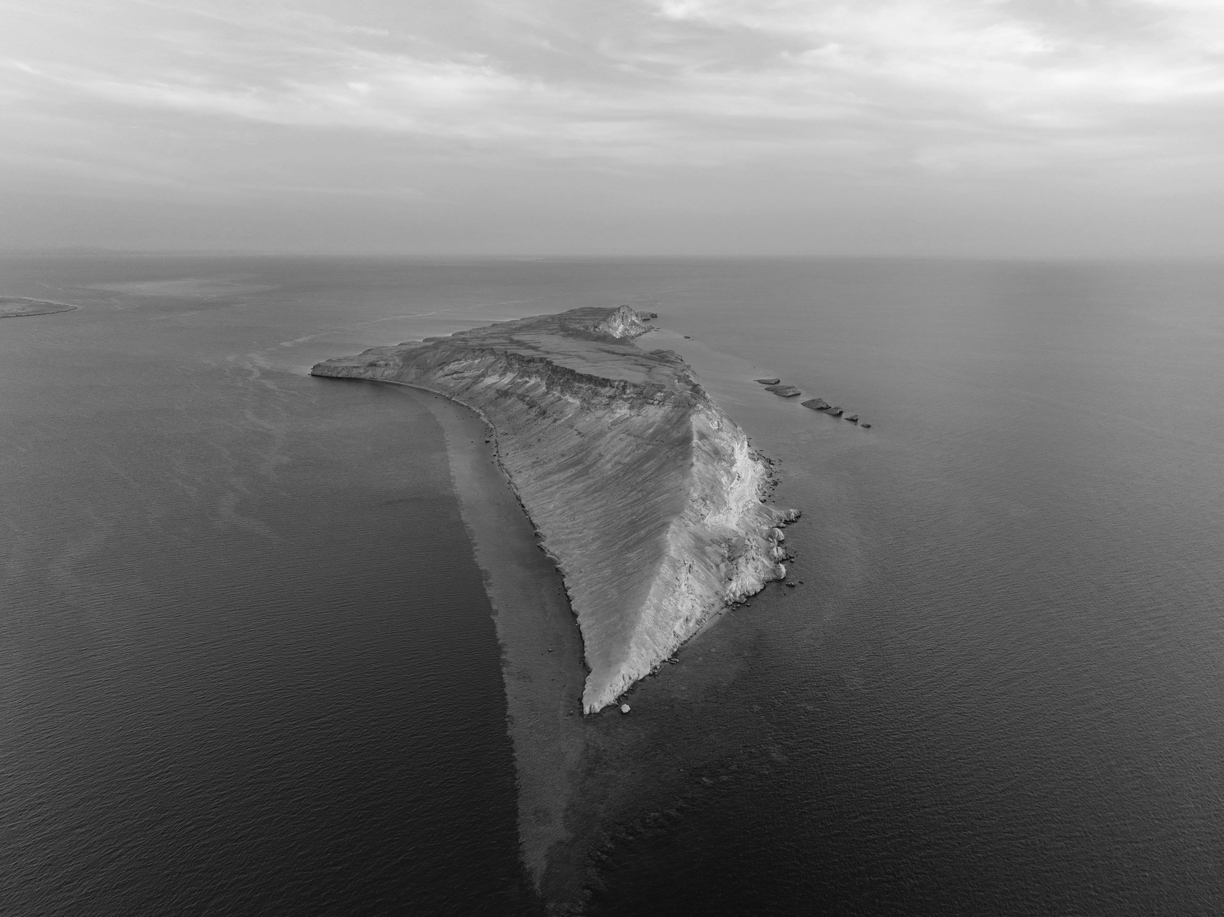 Grayscale aerial photograph of a long narrow rocky island surrounded by open sea, used as the original image for compression.